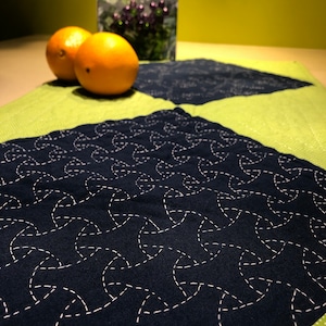 May include: Two bright orange oranges on a green and blue patterned fabric placemat. The fabric has a geometric design of white stitching on a dark blue background.