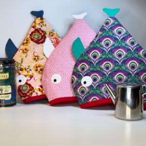 May include: Four whimsical fish-shaped tea cozies in various patterns and colors. One has a floral design, another is pink with white dots, and a third features a peacock feather pattern. Each cozy has a contrasting fin and eye details.