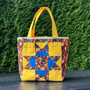 May include: A quilted tote bag with yellow handles and a star design. The star is made of yellow and blue fabric, with a seashell pattern on the sides. The bag has a zip closure and is designed for carrying items.