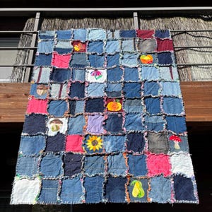 Könnte beinhalten: Eine Patchwork-Decke aus verschiedenen blauen Denim-Quadraten mit ausgefransten Rändern und eingearbeiteten Stoffflicken, die mit Motiven eines Ahornblatts, einer Blume, eines Kürbisses, einer Sonnenblume, eines Pilzes und einer Birne bestickt sind.