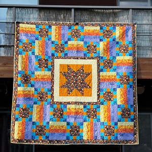 May include: A quilt with a star pattern in the center. The quilt is made of various fabrics in shades of orange, yellow, blue, and purple. The quilt is hanging on a wall.