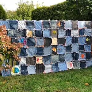 May include: A patchwork quilt made from denim fabric. The quilt is made up of squares of different shades of blue denim, with some squares featuring different patterns and designs. The quilt is hanging on a wooden fence in a garden setting.