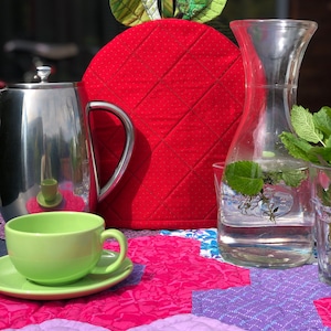 May include: A red quilted teapot cozy with green fabric leaves on top. The cozy is sitting on a colorful patchwork quilt with a green teacup and a glass pitcher of water with mint leaves.