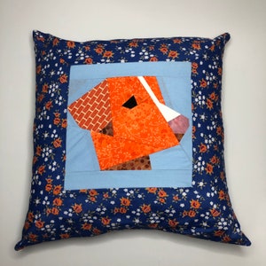 May include: A blue pillow with a floral pattern and a patchwork orange and brown dog face in the center.