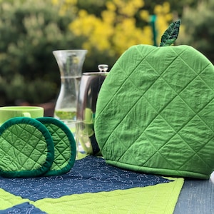 Könnte beinhalten: Zwei grüne, gesteppte Topflappen mit dunkelgrünem Rand und ein grüner, gesteppter Teekannenwärmer mit einem Blatt obenauf. Die Gegenstände liegen auf einem grünen und blauen, gesteppten Tischläufer.