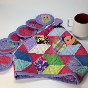 May include: A quilted tea cozy and matching coasters in a patchwork design. The cozy is primarily pink with various colorful fabric patches. The coasters are round with pink centers and lavender borders. A white mug with a red interior sits nearby.