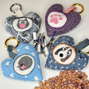 Puede incluir: Cuatro llaveros en forma de corazón con diseños de animales: un gato, una huella de pata, un perro y un erizo. Cada llavero tiene un lazo de tela y un anillo de metal. También hay pequeños pergaminos con cintas, junto con una dispersión de semillas marrones.