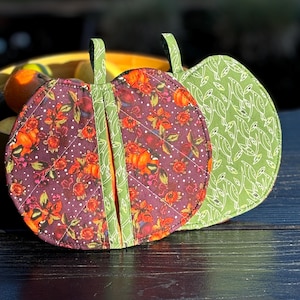 Può includere: Due presine trapuntate a forma di zucca. Una è di colore viola scuro con un motivo floreale e l'altra è di colore verde chiaro con un motivo a foglie.