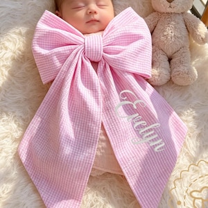 Può includere: Un neonato avvolto in una grande coperta rosa a forma di fiocco con il nome "Evelyn" ricamato in bianco. Un orsacchiotto beige è seduto accanto al bambino in una culla di legno con una soffice coperta bianca.