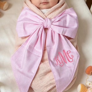 Könnte beinhalten: Ein Baby in einer rosa Wickeldecke mit einer großen rosa gestreiften Schleife. Die Schleife ist mit den Initialen "RMe" in Schreibschrift personalisiert. Im Hintergrund befinden sich zwei Teddybären und ein Plüschtier.