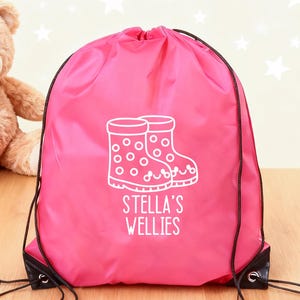 Puede incluir: Una bolsa con cordón rosa brillante con un gráfico blanco de botas de agua y el texto "STELLA'S WELLIES". La bolsa tiene cordones negros y esquinas reforzadas negras. Un oso de peluche está en el fondo.