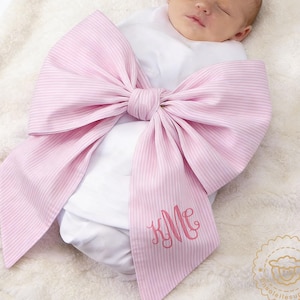 Peut inclure: Un nouveau-né emmailloté dans une couverture blanche et un grand nœud rose et blanc rayé. Le nœud porte un monogramme rose. Le bébé est allongé sur une couverture blanche moelleuse.