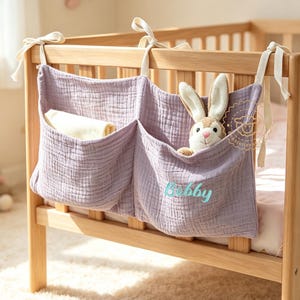 May include: A lavender muslin crib organizer with two pockets, tied to a wooden crib with fabric straps. One pocket holds a white cloth, and the other contains a plush rabbit toy. The name Bobby is embroidered in light blue on the front pocket.