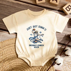 May include: Cream-colored baby onesie with the text "JUST GOT DUNK'D" and an illustration of a figure dunking a basketball. The text "BROOKLYN CHURCH 9.10.26" is below the illustration. Wooden blocks spell "BABY".