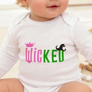 May include: A white baby bodysuit with long sleeves. The word "Wicked" is printed on the front in pink and green, with a pink crown above the word and a black witch hat to the right. The baby is sitting on a woven mat.