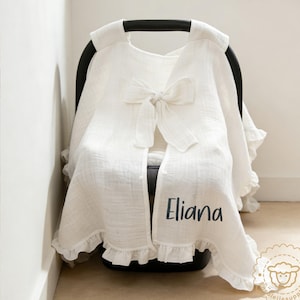 May include: White baby car seat cover with a large bow and ruffled edges. The cover is draped over a black car seat and features the name "Eliana" in black script. A logo with a sheep is visible.