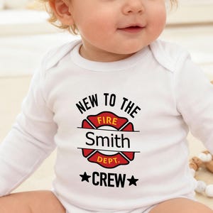 May include: A baby in a white long-sleeved onesie featuring a red and black firefighter emblem with the text "NEW TO THE FIRE DEPT. CREW" and a name. The baby is sitting on a light-coloured surface, with a neutral background.