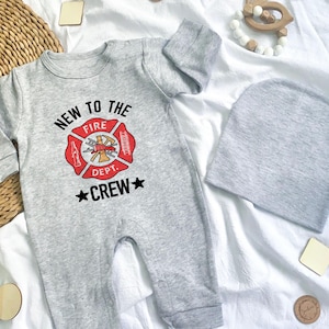 May include: A gray baby onesie and matching beanie set. The onesie features a red and gold firefighter emblem with the text "NEW TO THE FIRE DEPT. CREW" in black. The set is laid out on a white surface with wooden accessories.