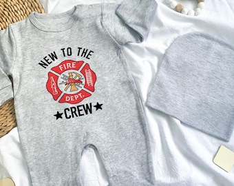 May include: A grey baby's onesie and beanie set. The onesie displays a red and gold firefighter emblem with the text "NEW TO THE FIRE DEPT. CREW" in black. The set is on a white surface with wooden accessories.