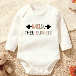 May include: White long-sleeve baby bodysuit with the text "MILK, THEN BURPEES" in black, brown, green, and orange. The text is above a barbell graphic. The bodysuit is surrounded by baby toys, including a teddy bear and a rainbow.