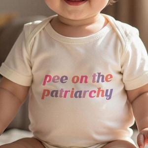 May include: A baby wearing a cream-coloured onesie with the text "pee on the patriarchy" in pink and purple. The baby has brown hair and is smiling. The background is a neutral colour.