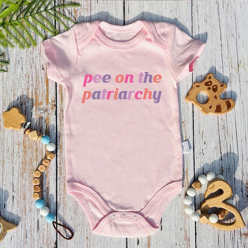 May include: A pale pink baby bodysuit with the slogan "pee on the patriarchy" in pink, orange, and purple. The image includes wooden baby toys and a pacifier clip with wooden beads.