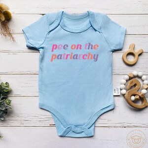 May include: Pale blue baby bodysuit featuring the text "pee on the patriarchy" in pink, orange, and purple. The bodysuit has short sleeves and popper fastenings. A wooden toy and a beaded teething ring are next to the bodysuit.