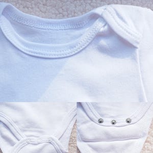 May include: Close-up of a white baby onesie with the text "cloth details". The onesie has a round neckline, snap closures at the bottom, and detailed stitching. The fabric looks soft and comfortable.
