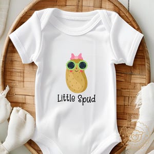 May include: White baby bodysuit with a cartoon potato wearing sunglasses and a pink bow. The text "Little Spud" is printed below the potato graphic. The bodysuit is made of soft material.