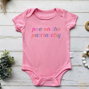 May include: A pink baby bodysuit with short sleeves. The text "pee on the patriarchy" is printed across the front in a gradient of pink, orange, and purple. The bodysuit has a round neckline and snaps at the bottom.