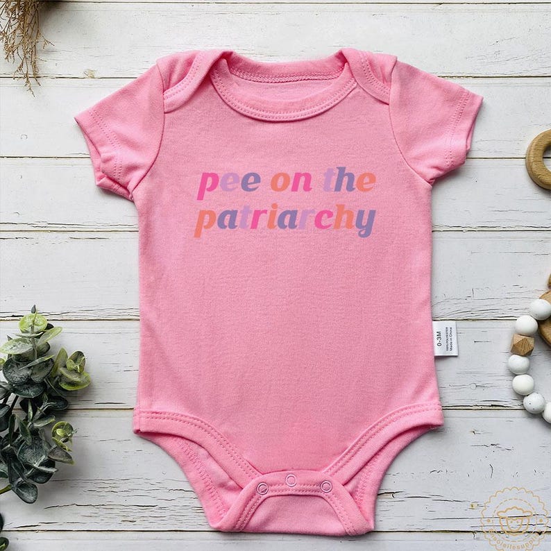 May include: A pink baby bodysuit with short sleeves. The text "pee on the patriarchy" is printed across the front in a gradient of pink, orange, and purple. The bodysuit has a round neckline and snaps at the bottom.