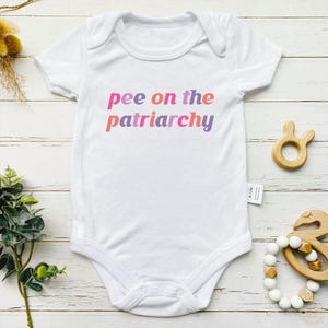 May include: White baby bodysuit with the text "pee on the patriarchy" in pink, orange, and purple. The short-sleeved bodysuit has a snap closure. Wooden baby toys are also visible in the image.