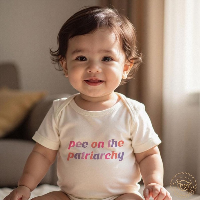 May include: A baby wearing a cream-coloured onesie with the text "pee on the patriarchy" in pink and purple. The baby has brown hair and is smiling. The background is a neutral colour.