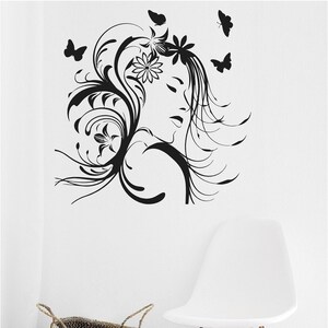Pu&ograve; includere: Decorazione murale in bianco e nero raffigurante un volto femminile con dettagli floreali e farfalle. Una sedia bianca con gambe in legno e un cesto intrecciato con una coperta grigia sono in primo piano.