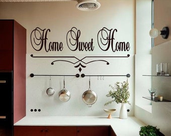 Puede incluir: Una cocina blanca con un estante de metal negro para ollas y tres ollas plateadas colgadas. La pared detrás del estante tiene una calcomanía de vinilo negro que dice "Home Sweet Home" con un adorno decorativo.