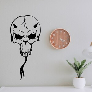 May include: Black and white skull graphic with a long, thin tail, on a white wall with a wooden clock and a potted plant.