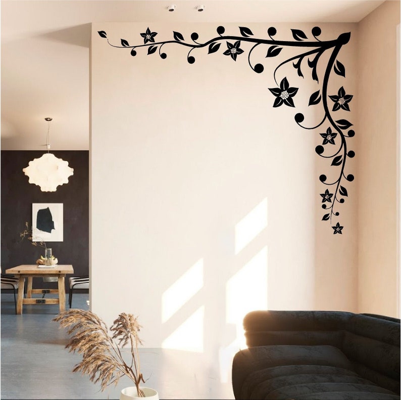Puede incluir: Pegatina de pared floral negra con un dise&ntilde;o de vid. La pegatina est&aacute; en la esquina de una habitaci&oacute;n con una pared blanca y un sof&aacute; en primer plano.