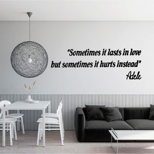 May include: A black and white wall decal with a quote by Adele: "Sometimes it lasts in love but sometimes it hurts instead." The quote is displayed on a white wall in a living room setting with a black sofa and a white dining table with chairs.