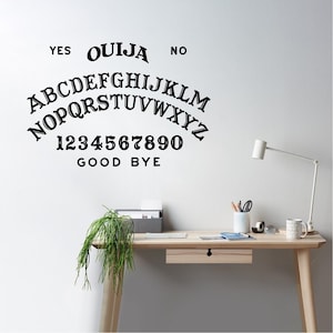 Puede incluir: Calcomanía de pared negra de la tabla Ouija con las palabras "YES", "NO" y "GOOD BYE". La calcomanía también incluye el alfabeto y los números 0-9. La calcomanía se muestra en una pared blanca encima de un escritorio de madera.