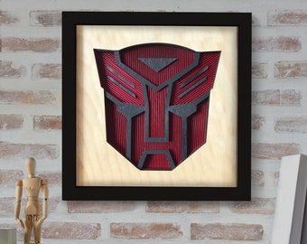 May include: A black framed wall art piece with a red and black 3D cutout of the Autobot symbol from the Transformers franchise.