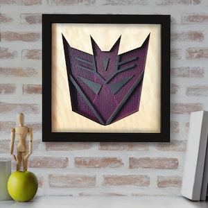 May include: A black framed wall art piece with a purple and black 3D cutout of the Decepticon symbol from the Transformers franchise.