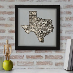 May include: A framed 3D wood map of Texas with a detailed outline of the state's roads and highways. The map is mounted on a gray background and framed in black.