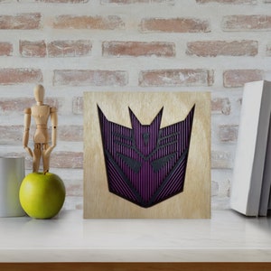 May include: Wooden wall art featuring a purple and black geometric design. The artwork is mounted on a light-colored wooden square. The design has a layered effect, with the purple elements creating depth. The piece is displayed on a white surface.