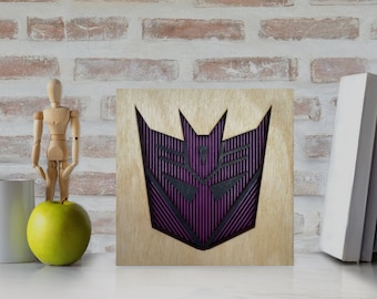 May include: Wooden wall art featuring a purple and black geometric design. The artwork is mounted on a light-colored wooden square. The design has a layered effect, with the purple elements creating depth. The piece is displayed on a white surface.