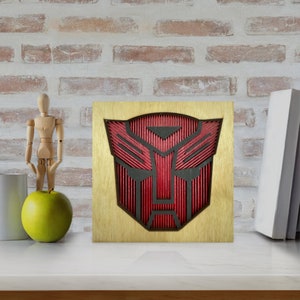 May include: A wooden wall art piece featuring the Transformers logo in red and black. The logo is cut out of the wood and has a 3D effect.