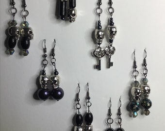 May include: Six pairs of silver skull earrings with black beads and charms. The earrings are on a white background.