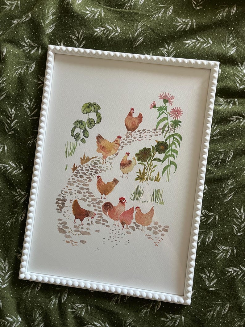 Op de afbeelding: Een ingelijst aquarel schilderij van een groep kippen die over een stenen pad lopen. De kippen zijn bruin en wit met rode kammen. Het pad is omgeven door groene planten en roze bloemen.