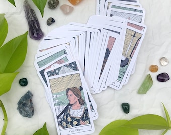Puede incluir: Una lectura de cartas de tarot con un despliegue de cartas sobre una superficie blanca. La carta del centro es El Loco, que muestra a una persona con una mochila y un perro caminando hacia un acantilado. La carta está rodeada de hojas verdes y cristales.