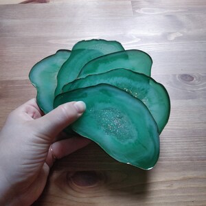 May include: A collection of five green agate coasters with gold edging. Each coaster features a geode-style centre with glitter. The coasters are fanned out, highlighting their unique design and colour.