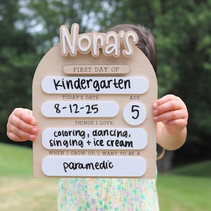 Pu&ograve; includere: Un cartello di legno con il nome "Nora's" in alto e le parole "First Day of Kindergarten" sotto. Il cartello include la data "8-12-25", l'et&agrave; "5" ed elenca "coloring, dancing, singing + ice cream" come cose amate. Dice anche "paramedic".